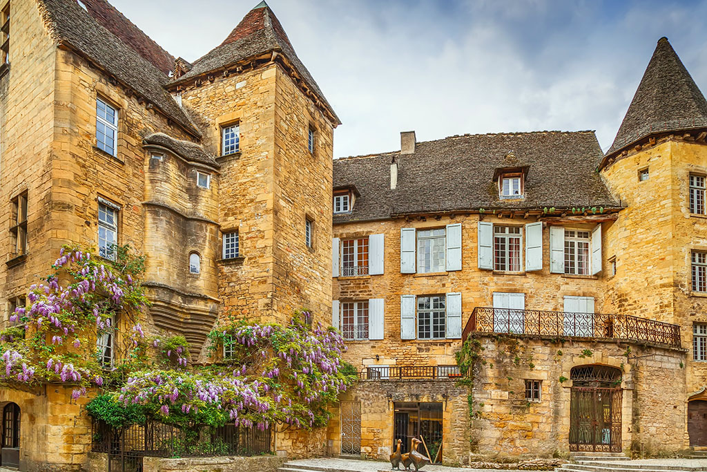 A beautiful medieval building in Sarlat, France, with a stone facade, a slate roof, and a balcony adorned with purple flowers. This is one of the best places to stay in Sarlat for a luxurious experience.