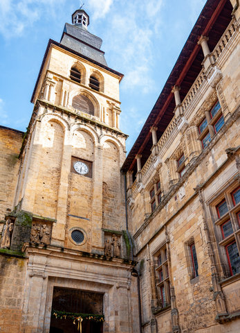A tall, stone bell tower with ornate detailing, surrounded by other historic buildings in Sarlat-la-Canéda, France.